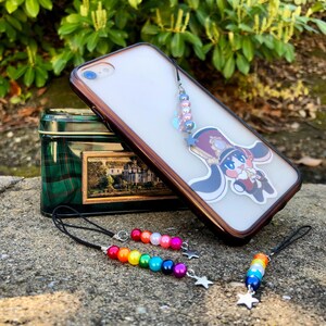 May include: A clear phone case with a brown border and a cartoon character design. The case has a beaded charm with a star pendant. Additional beaded charms in rainbow colors are also displayed, along with a green and gold tin.