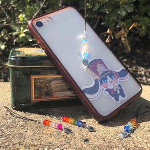May include: A clear phone case with a maroon border, featuring a cartoon character sticker and a dangling charm with colorful beads and heart-shaped charms. A decorative tin and additional beaded charms are also visible.