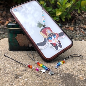 May include: Clear phone case with a brown border, featuring a cartoon character charm and a beaded charm. The beaded charms include rainbow and clear beads with star-shaped charms. A green tin is in the background.