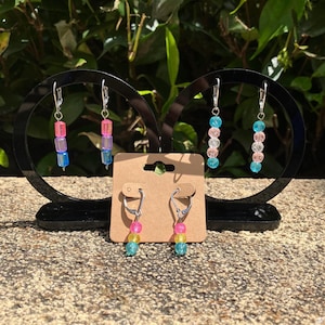 May include: Three pairs of dangle earrings with colourful beads. The earrings are displayed on a black earring stand. The beads are arranged in the colours of the transgender, bisexual, and pansexual pride flags.