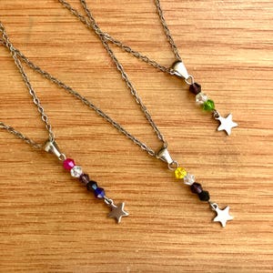 Könnte beinhalten: Drei silberne Halsketten mit farbigen Perlenanhängern und Sternanhängern. Jede Halskette hat ein anderes Farbschema: rosa, lila und blau; gelb, klar und lila; und grün, klar und lila. Die Halsketten werden auf einer Holzoberfläche präsentiert.