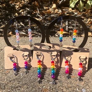 May include: Earrings with colorful beads in rainbow hues and pastel shades. Each earring features a series of beads, suspended from a silver-tone hook, and a small star charm. The earrings are displayed on small cardboard tags.