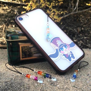 May include: A clear phone case with a brown wooden frame, featuring a cartoon character sticker and a beaded charm. Additional beaded charms with heart-shaped pendants are also visible, along with a decorative tin.