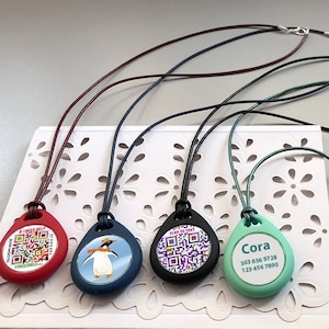 May include: A collection of colorful pendant necklaces with various designs. The pendants are red, blue, black, and mint green, each with a different graphic or text. The necklaces have black, brown, and green cords with silver clasps.
