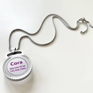 May include: Silver necklace featuring a circular pendant with the name "Cora" and two phone numbers in purple text. The pendant has a clear face. The chain is a fine, linked design with an adjustable clasp.
