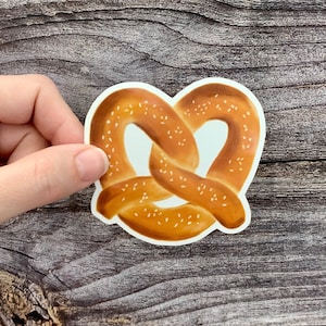 May include: A brown pretzel with white sesame seeds. The pretzel is shaped like a knot and is on a white background.
