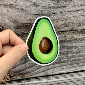 May include: A green avocado sticker with a brown pit. The sticker is on a wooden background.