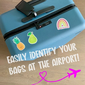 May include: A blue suitcase with stickers on it. The stickers include a pineapple, a pear, a rainbow with the text "Choose kindness", and the text "EASILY IDENTIFY YOUR BAGS AT THE AIRPORT!" with a pink heart and airplane graphic.