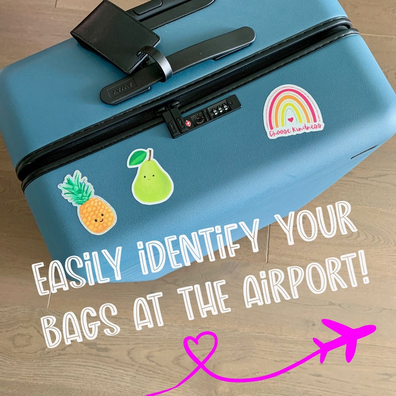 May include: A blue suitcase with stickers on it. The stickers include a pineapple, a pear, a rainbow with the text "Choose kindness", and the text "EASILY IDENTIFY YOUR BAGS AT THE AIRPORT!" with a pink heart and airplane graphic.