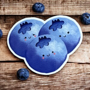 May include: Three blue cartoon blueberries with happy faces and white eyes. The blueberries are arranged in a cluster on a wooden surface.