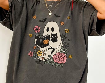 Può includere: T-shirt grigio scuro con un fantasma che legge un libro con una zucca, un gatto nero, fiori e pipistrelli. Il design è circondato da fiori e foglie colorate. La maglietta ha maniche corte e una vestibilità ampia.