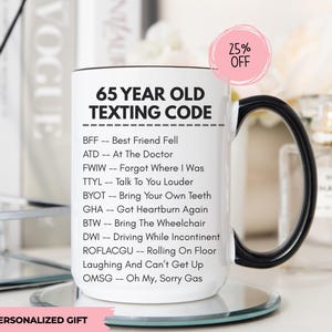 May include: White ceramic mug with a black handle and rim, featuring the text "65 YEAR OLD TEXTING CODE" and humorous abbreviations. A pink circle with "25% OFF" is visible. The mug sits on a mirrored surface. The words "PERSONALIZED GIFT" are at the bottom.