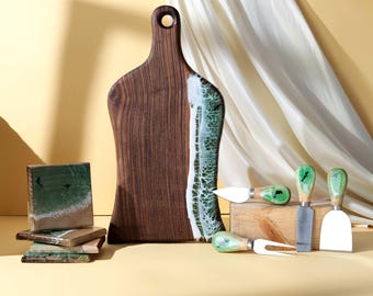 May include: A dark wooden serving board with a green and white resin edge, alongside cheese knives and coasters. The set is designed for serving, with a natural aesthetic and a touch of coastal design.