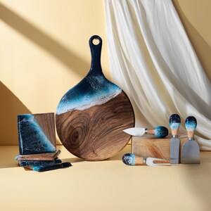 Puede incluir: Una tabla de servir de madera con una incrustación de resina azul y blanca que representa olas del océano. La tabla tiene un mango y viene acompañada de cuatro pequeños cuchillos para queso con mangos de resina azul y blanca.