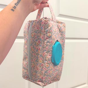 May include: A floral print wipe dispenser bag with a teal blue lid. The bag has a zipper closure and a handle. The floral pattern includes pink, blue, and yellow flowers on a light pink background.