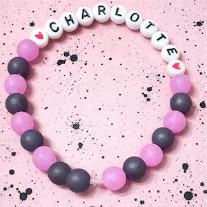 以下が含まれることがあります： 白い文字ビーズで「CHARLOTTE」と綴られたビーズブレスレット。濃いグレーと薄いピンクのビーズが交互に並び、ハート型のアクセントが付いています。