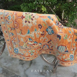 Puede incluir: Una alfombra naranja estampada con un diseño detallado, colgada sobre el manillar de una bicicleta vintage. La alfombra presenta una variedad de colores y elementos florales. La palabra "FAIRFAX" está impresa en la alfombra.