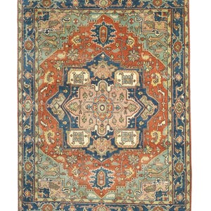 Puede incluir: Alfombra rectangular con un diseño intrincado y simétrico. La alfombra presenta un medallón central en tonos rosa y beige, rodeado por un patrón de azul, óxido y verde. El borde es azul oscuro con motivos florales.