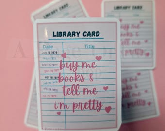 May include: A collection of stickers with a library card design in pink and white. The stickers display the text "buy me books & tell me I'm pretty" in pink script, decorated with small pink hearts. The background is a pale pink.