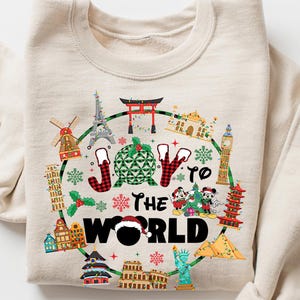 Puede incluir: Sudadera beige con un diseño circular que presenta monumentos como la Torre Eiffel y el Big Ben, y el texto "JOY TO THE WORLD". El diseño incluye elementos navideños y personajes de dibujos animados.