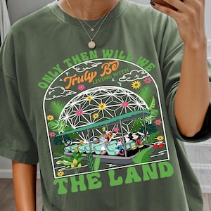 Könnte beinhalten: Olivgrünes T-Shirt mit einer Grafik, die eine Themenpark-Fahrt und den Text "ONLY THEN WILL WE TRULY BE LIVING WITH THE LAND" zeigt. Das Design beinhaltet farbenfrohe Illustrationen von Charakteren und floralen Elementen.