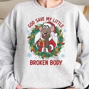 May include: A light gray sweatshirt featuring a cartoon rat wearing a Santa hat and holding a gift. The rat is surrounded by a wreath with the text "GOD SAVE MY LITTLE BROKEN BODY" in red. Blue snowflakes are scattered around the design.
