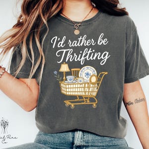 May include: Dark gray t-shirt with the text "I'd rather be Thrifting" in white script. The shirt features a graphic of a shopping cart filled with vintage items like a lamp, dishes, and a teacup. The shirt is made of a soft, comfortable material.