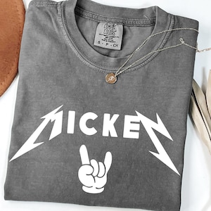 May include: A gray Comfort Colors t-shirt with the word "MICKEY" in a white, stylized font, and a white hand gesture. A silver necklace with a pendant is on the shirt.