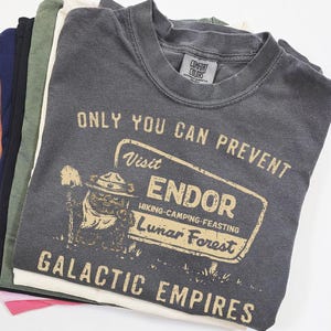 Puede incluir: Una pila de camisetas de varios colores. La camiseta superior es de color gris oscuro y tiene el texto "ONLY YOU CAN PREVENT Visit ENDOR Hiking-Camping-Feasting Lunar Forest GALACTIC EMPIRES". Las camisetas están dobladas y apiladas.