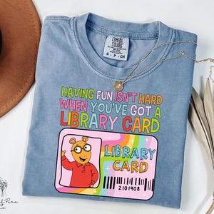 May include: A blue Comfort Colors t-shirt with the text "HAVING FUN ISN'T HARD WHEN YOU'VE GOT A LIBRARY CARD." A library card graphic features a cartoon character and a barcode. A gold necklace is draped across the shirt.