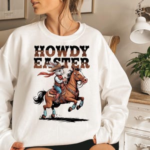 May include: White sweatshirt with the text "HOWDY EASTER" in brown letters. The graphic depicts a cartoon rabbit wearing a cowboy hat and riding a brown horse. The rabbit is wearing a red scarf, blue jeans, and boots. The horse is jumping.