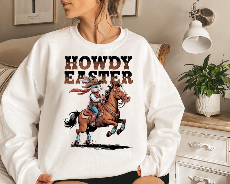 May include: White sweatshirt with the text "HOWDY EASTER" in brown letters. The graphic depicts a cartoon rabbit wearing a cowboy hat and riding a brown horse. The rabbit is wearing a red scarf, blue jeans, and boots. The horse is jumping.