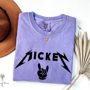 May include: A lilac-colored t-shirt with the word "MICKEY" in a black, stylized font, and a black hand gesture. The shirt has a crew neck and short sleeves. A gold necklace with a pendant is on the shirt.