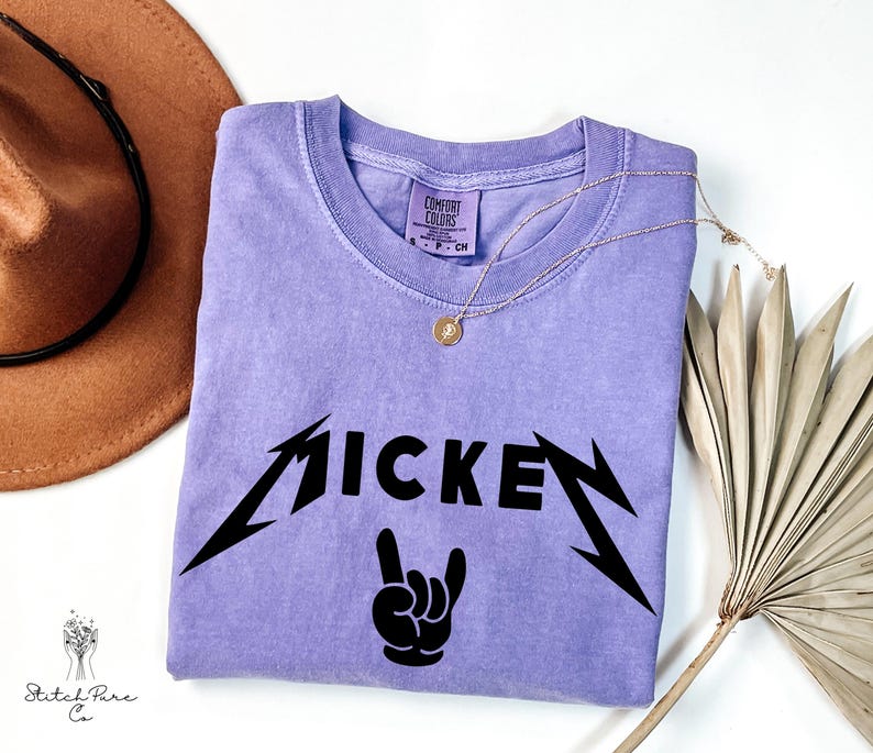 May include: A lilac-colored t-shirt with the word "MICKEY" in a black, stylized font, and a black hand gesture. The shirt has a crew neck and short sleeves. A gold necklace with a pendant is on the shirt.