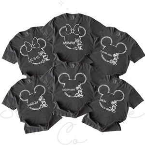 May include: A collection of dark gray t-shirts featuring white Mickey Mouse designs. Some shirts have text like "Mommy", "Lil Sis", "Daddy", "Back", and "Custom Name". The shirts are likely for a family or group.