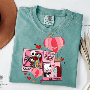 Puede incluir: Una camiseta Comfort Colors de color verde azulado con un dise&ntilde;o gr&aacute;fico con personajes de Pesadilla antes de Navidad. El dise&ntilde;o incluye globos aerost&aacute;ticos rosas, corazones y las frases "XOXO" y "BE MINE". Un collar plateado con un colgante de coraz&oacute;n est&aacute; sobre la camiseta.