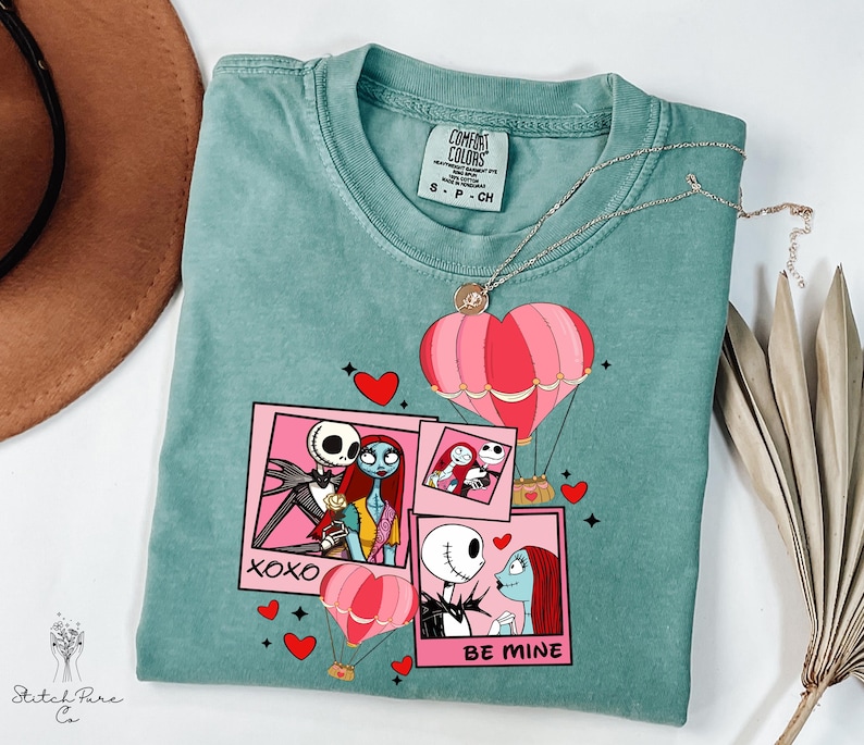 Puede incluir: Una camiseta Comfort Colors de color verde azulado con un dise&ntilde;o gr&aacute;fico con personajes de Pesadilla antes de Navidad. El dise&ntilde;o incluye globos aerost&aacute;ticos rosas, corazones y las frases "XOXO" y "BE MINE". Un collar plateado con un colgante de coraz&oacute;n est&aacute; sobre la camiseta.