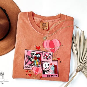 Puede incluir: Una camiseta de color coral con un dise&ntilde;o gr&aacute;fico con im&aacute;genes de Jack y Sally de The Nightmare Before Christmas. El dise&ntilde;o incluye globos aerost&aacute;ticos rosas, corazones y las frases "XOXO" y "BE MINE."