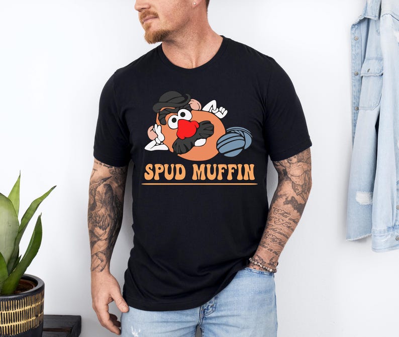May include: Black t-shirt featuring a cartoon character with a hat, red nose, and blue gloves. The text "SPUD MUFFIN" is printed in orange letters below the character. The shirt is made of a soft material.