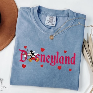 Op de afbeelding: Een lichtblauw t-shirt met het woord "Disneyland" in roze script, met een cartoon Mickey Mouse als Cupido met pijl en boog. Rode harten zijn verspreid rond de tekst. Een gouden ketting is over het shirt gedrapeerd.