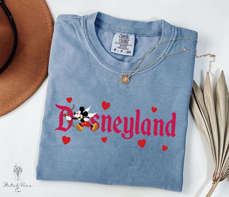 Op de afbeelding: Een lichtblauw t-shirt met het woord "Disneyland" in roze script, met een cartoon Mickey Mouse als Cupido met pijl en boog. Rode harten zijn verspreid rond de tekst. Een gouden ketting is over het shirt gedrapeerd.