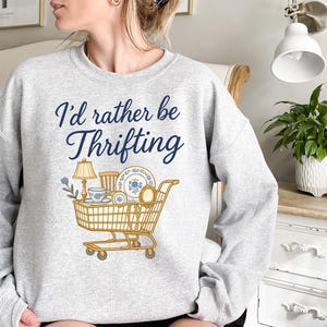 May include: Light gray sweatshirt with the text "I'd rather be Thrifting" in navy blue script. A shopping cart graphic is filled with thrifted items, including a lamp, teacup, and plates.