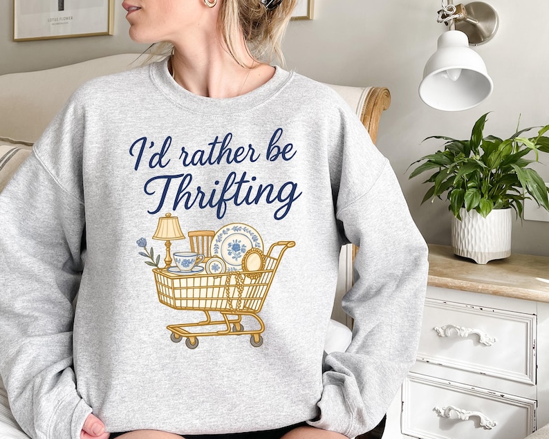 May include: Light gray sweatshirt with the text "I'd rather be Thrifting" in navy blue script. A shopping cart graphic is filled with thrifted items, including a lamp, teacup, and plates.