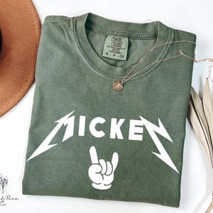 May include: Olive green t-shirt with the word "MICKEY" in a white, stylized font, and a white hand gesture. A gold necklace with a pendant rests on the shirt. The shirt is folded on a white surface.