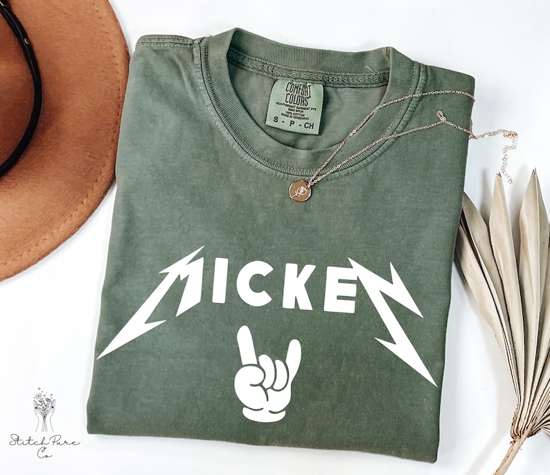 May include: Olive green t-shirt with the word "MICKEY" in a white, stylized font, and a white hand gesture. A gold necklace with a pendant rests on the shirt. The shirt is folded on a white surface.