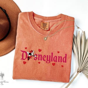 Op de afbeelding: Koraalrood T-shirt met het woord "Disneyland" in roze script, met een Minnie Mouse-afbeelding met Cupido-vleugels en rode harten. Een gouden ketting en een bruine hoed zijn ook zichtbaar.