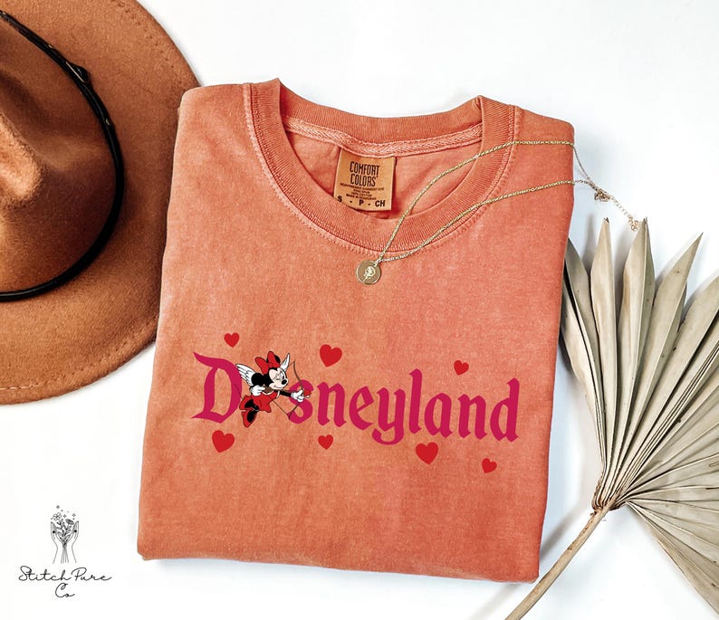 Op de afbeelding: Koraalrood T-shirt met het woord "Disneyland" in roze script, met een Minnie Mouse-afbeelding met Cupido-vleugels en rode harten. Een gouden ketting en een bruine hoed zijn ook zichtbaar.