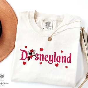 Op de afbeelding: Cr&egrave;mekleurig T-shirt met het woord "Disneyland" in roze script, met een Minnie Mouse-afbeelding en rode harten. Een gouden ketting en een bruine hoed zijn zichtbaar op de achtergrond. Het shirt is van Comfort Colors.