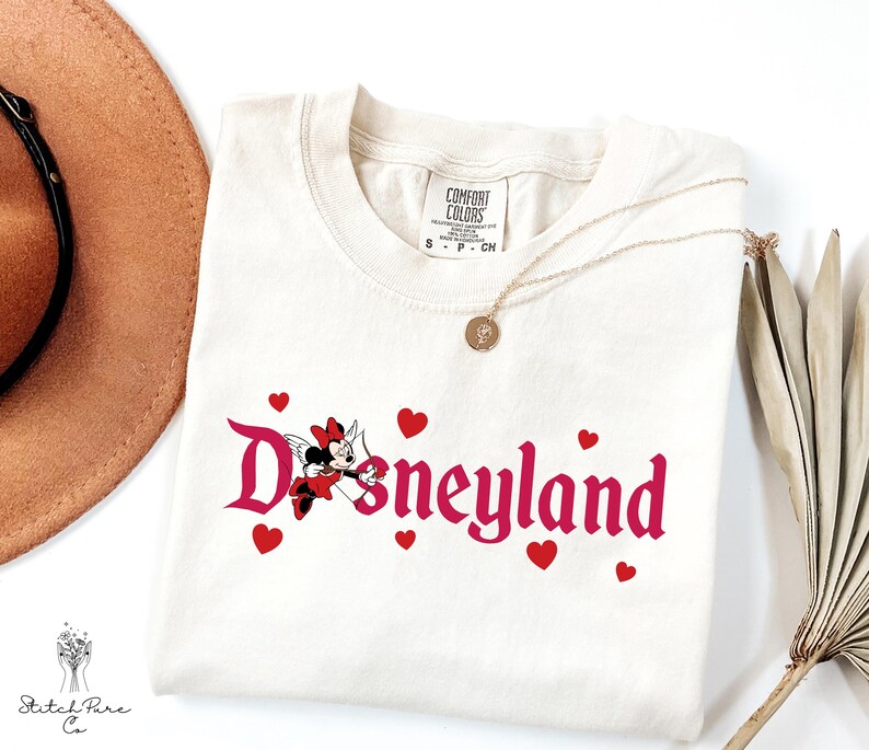 Op de afbeelding: Cr&egrave;mekleurig T-shirt met het woord "Disneyland" in roze script, met een Minnie Mouse-afbeelding en rode harten. Een gouden ketting en een bruine hoed zijn zichtbaar op de achtergrond. Het shirt is van Comfort Colors.