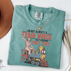 May include: A teal Comfort Colors t-shirt with the text "I'M NOT ALWAYS A TRAIN WRECK JUST KIDDING TOOT TOOT" and a graphic of a woman on a train. A gold necklace is draped across the shirt. The shirt is folded on a white surface.